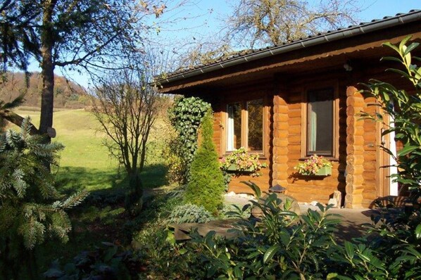 A rustic log cabin with a stone foundation, surrounded by a well-maintained garden and a view of a grassy field.