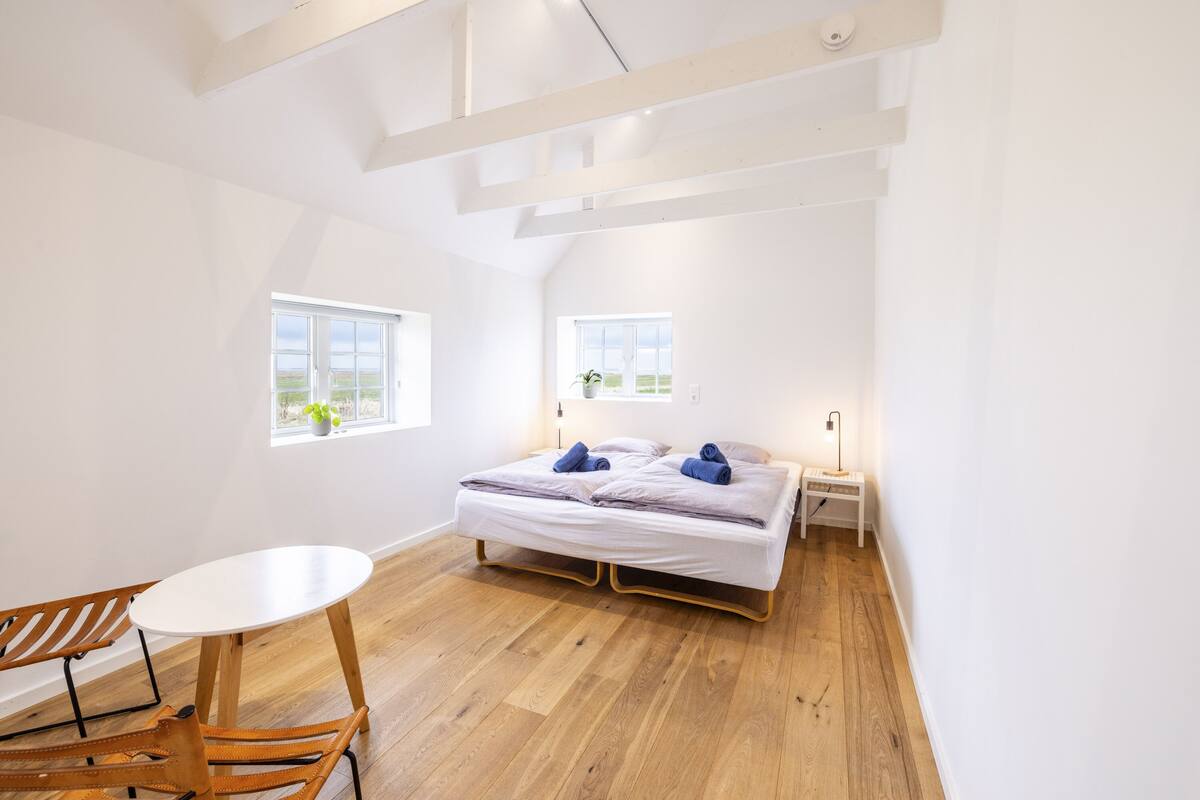 Un dormitorio con cama, mesita de noche, lámpara, ventana y mesa de comedor con sillas.