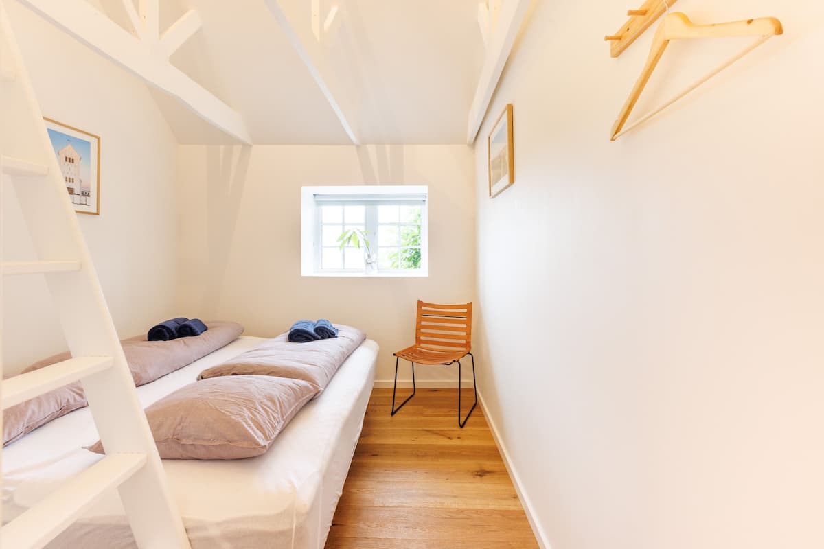Una habitación pequeña y luminosa con una cama individual, una silla de madera y un ventanal que deja entrar luz natural.
