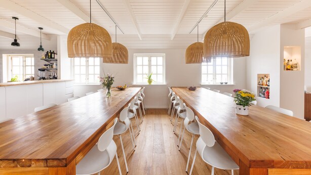 Une cuisine spacieuse avec une longue table en bois, des chaises blanches, des suspensions en osier et une fenêtre offrant une belle vue.