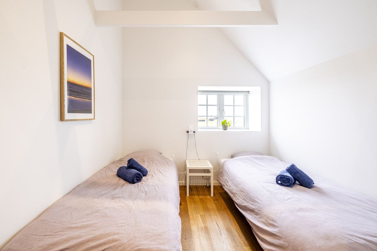 Una habitación pequeña y luminosa con dos camas individuales, una ventana y un cuadro en la pared.