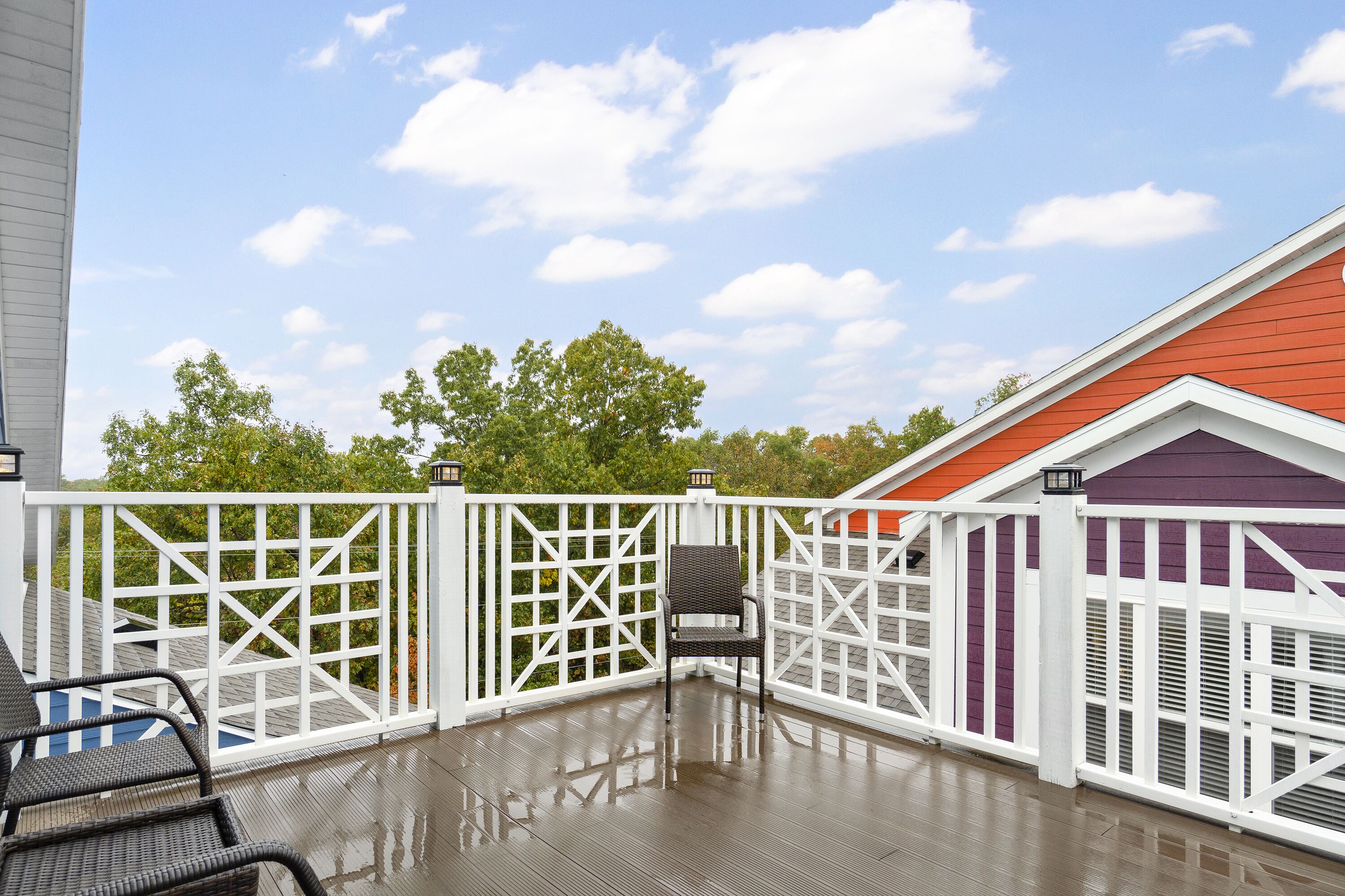 Une terrasse extérieure couverte, agrémentée d'une clôture en treillis blanche, d'un fauteuil en osier noir et d'un bâtiment violet en arrière-plan.