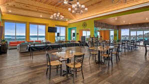 Une salle à manger spacieuse, dotée de tables et de chaises en bois, de grandes fenêtres offrant une vue sur le paysage, et d’un plafond orné de poutres en bois et de lustres.
