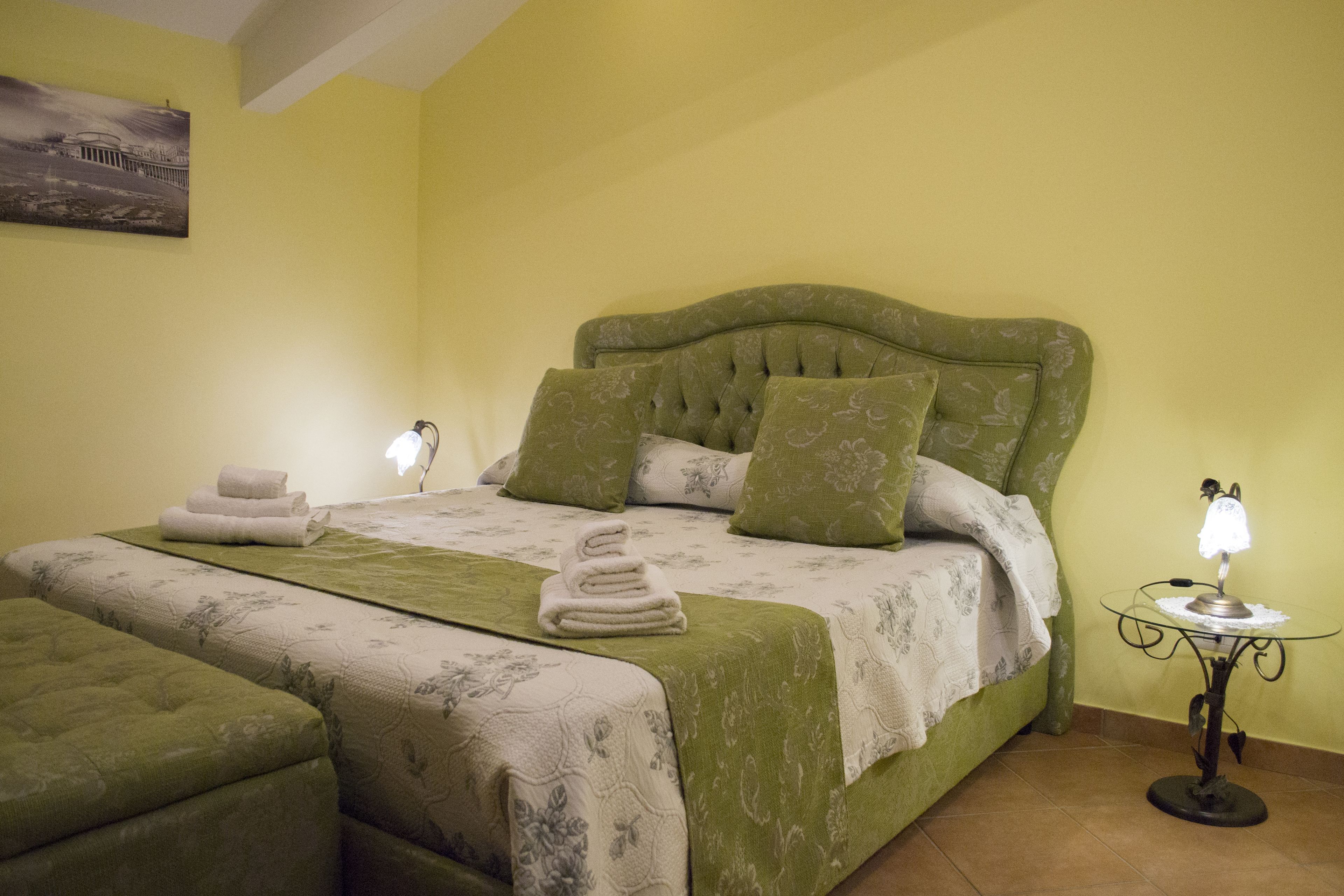 A neatly made bed with green bedding and pillows, a bedside table with a lamp, and a wall-mounted picture.