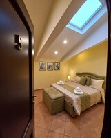 Een slaapkamer met dakraam, een bed met kussens en dekbed, een nachtkastje met lamp en een deur met nummer.