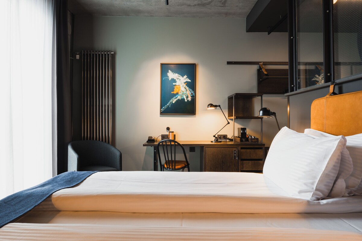 A hotel room with a bed, a desk, a chair, a lamp, and a painting on the wall.