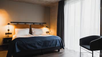 Una habitación de hotel moderna con una cama grande, mesitas de noche con lámparas, una silla negra y un amplio ventanal con cortinas.