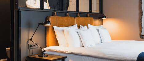 Una cama bien hecha con ropa de cama blanca y un cabecero con un espejo circular. Se ve una mesita de noche con una lámpara.