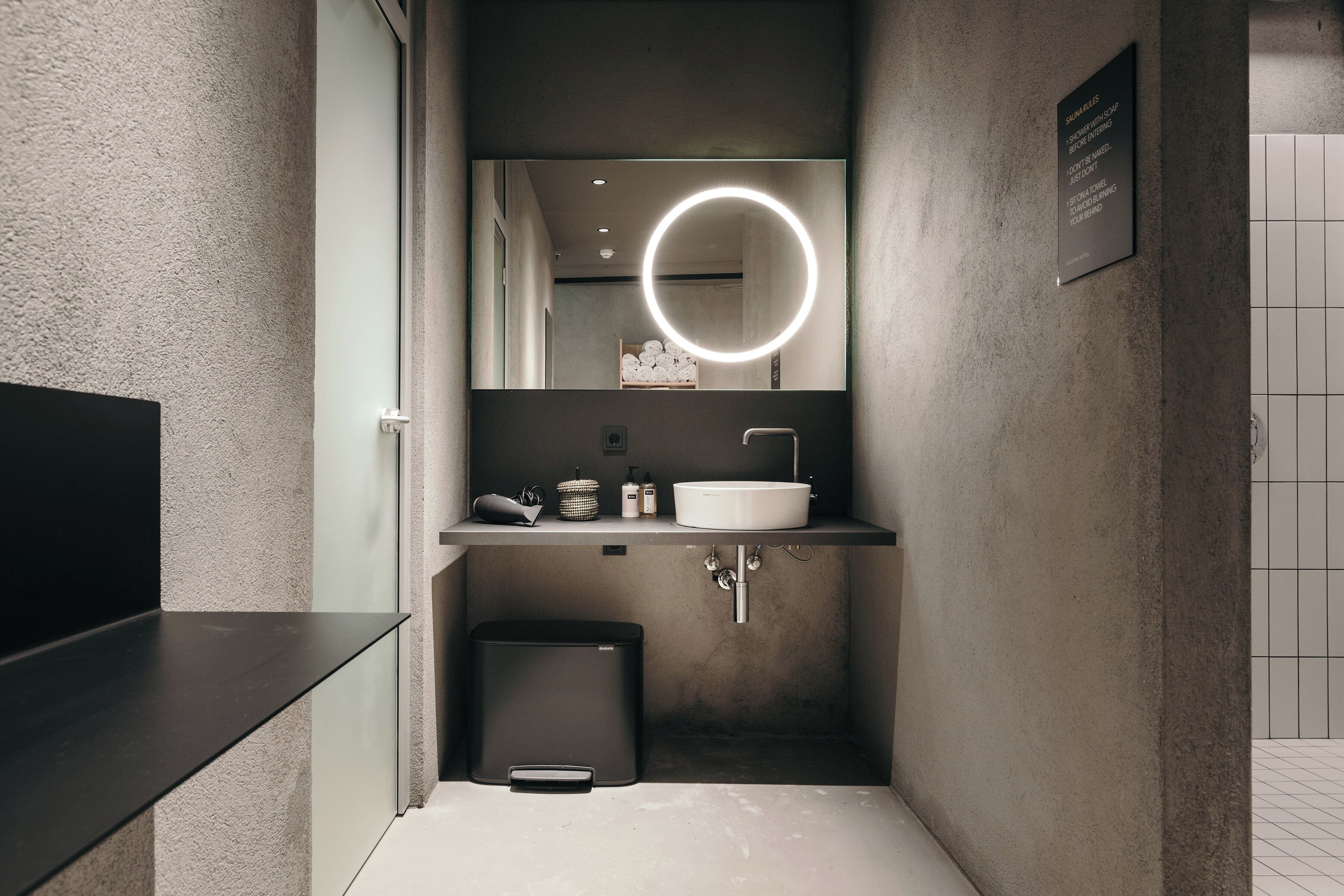 A modern bathroom with a large mirror, a round light, a sink, and a trash bin.
