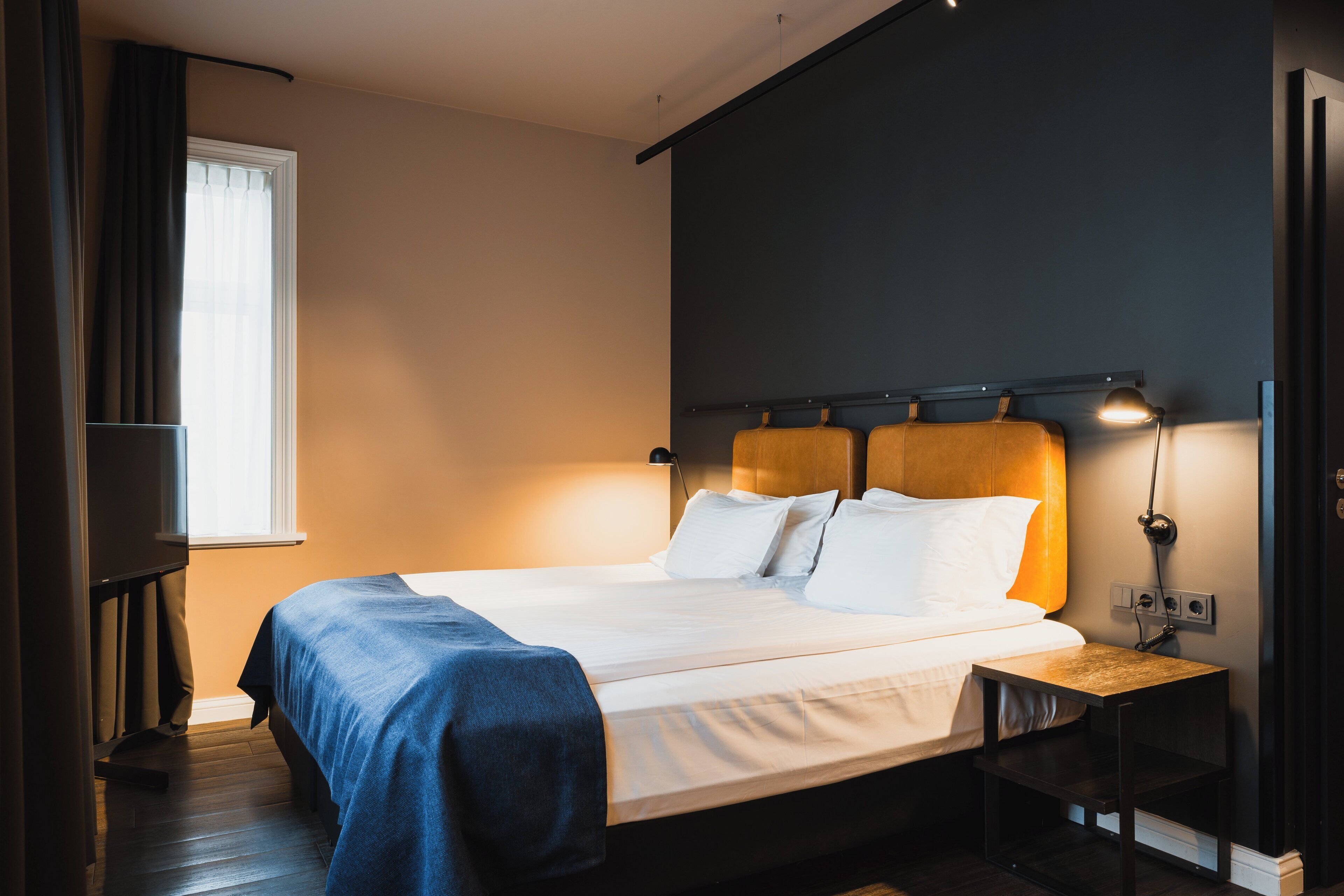 Un lit bien fait, avec du linge de lit blanc et une couverture bleue, une table de chevet avec une lampe, et une fenêtre avec des rideaux.