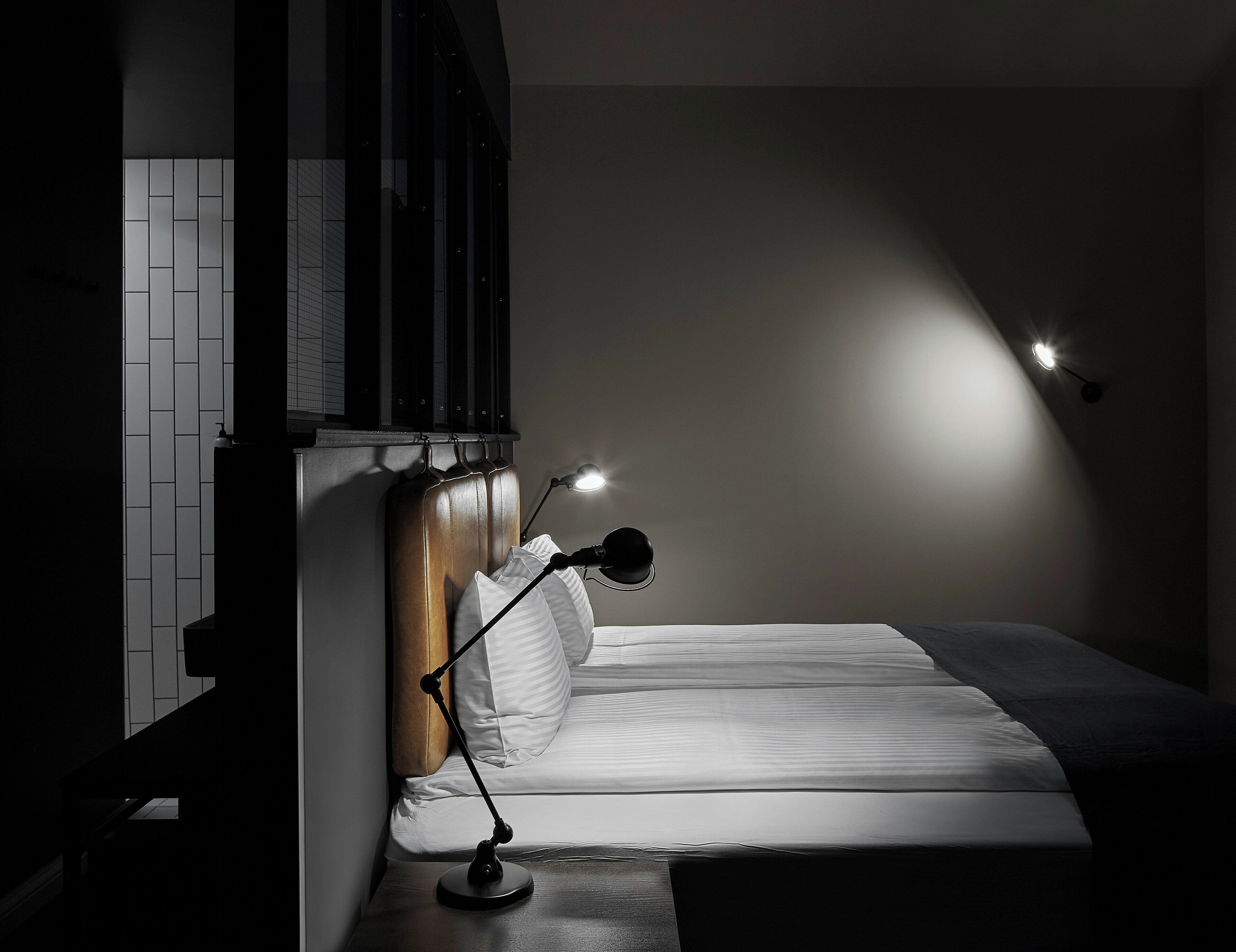 A bedroom with a bed, a bedside lamp, a wall-mounted shelf, and a window with blinds.
