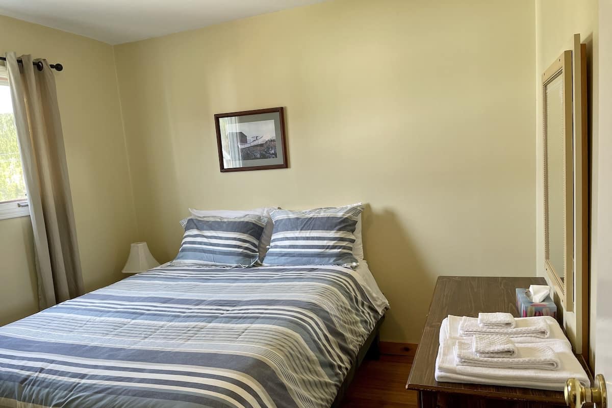 A neatly made bed with striped bedding, a bedside table with towels, a lamp, and a framed picture on the wall.