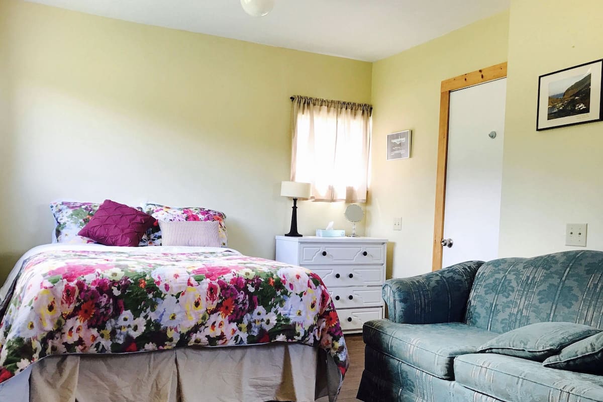A bedroom with a floral bedspread, a bedside table with a lamp, a dresser, and a green sofa.