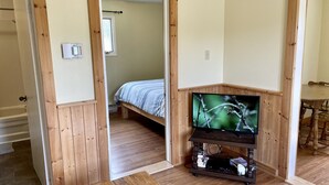 A room with a wooden floor, a bed with a striped bedspread, a television on a wooden stand, and a dining table with chairs.