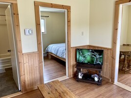 Una habitación con piso de madera, una cama con colcha a rayas, un televisor sobre un mueble de madera y una mesa de comedor con sillas.