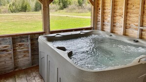 A wooden cabin with a hot tub and a view of a green field and trees.