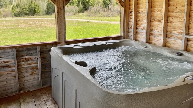 A wooden cabin with a hot tub and a view of a green field and trees.