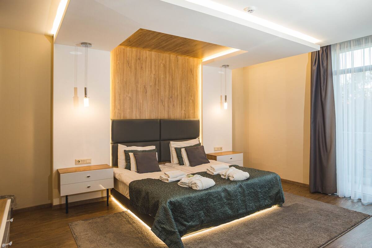 Ein modernes Schlafzimmer mit einem großen Bett, Nachttischen, einer Wand im Holzoptik und Blick ins Freie durch transparente Vorhänge.