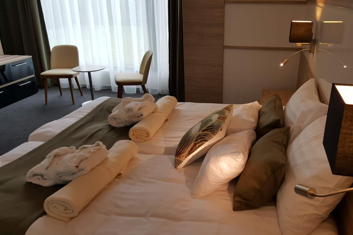 Ein ordentlich bezogenes Bett mit aufgerollten Handtüchern, ein Kissen mit Blättermotiv und eine Nachttischlampe.