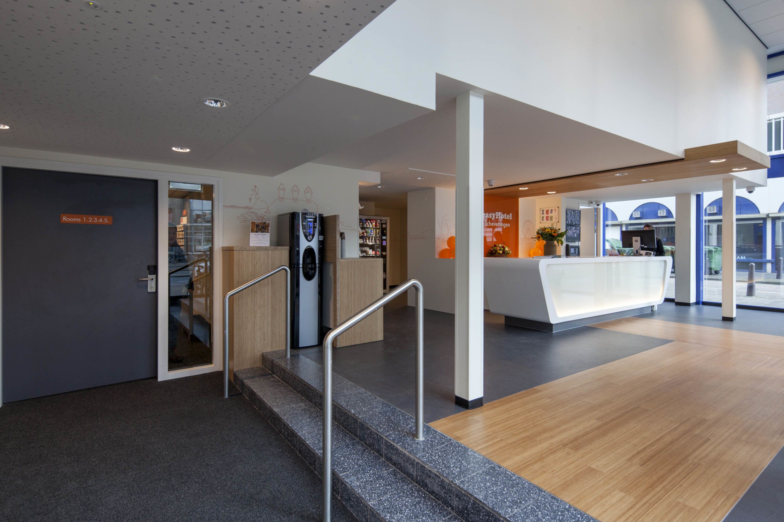 Un hall d’hôtel moderne avec un comptoir d’accueil, un escalier doté de rampes en métal et un panneau indiquant les numéros de chambre : 1-2, 3-4, 5.