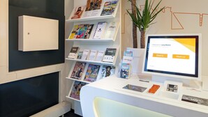 Een receptiebalie met een computer beeldscherm waarop een incheckinterface staat, een plant en een wandplank met tijdschriften en brochures.