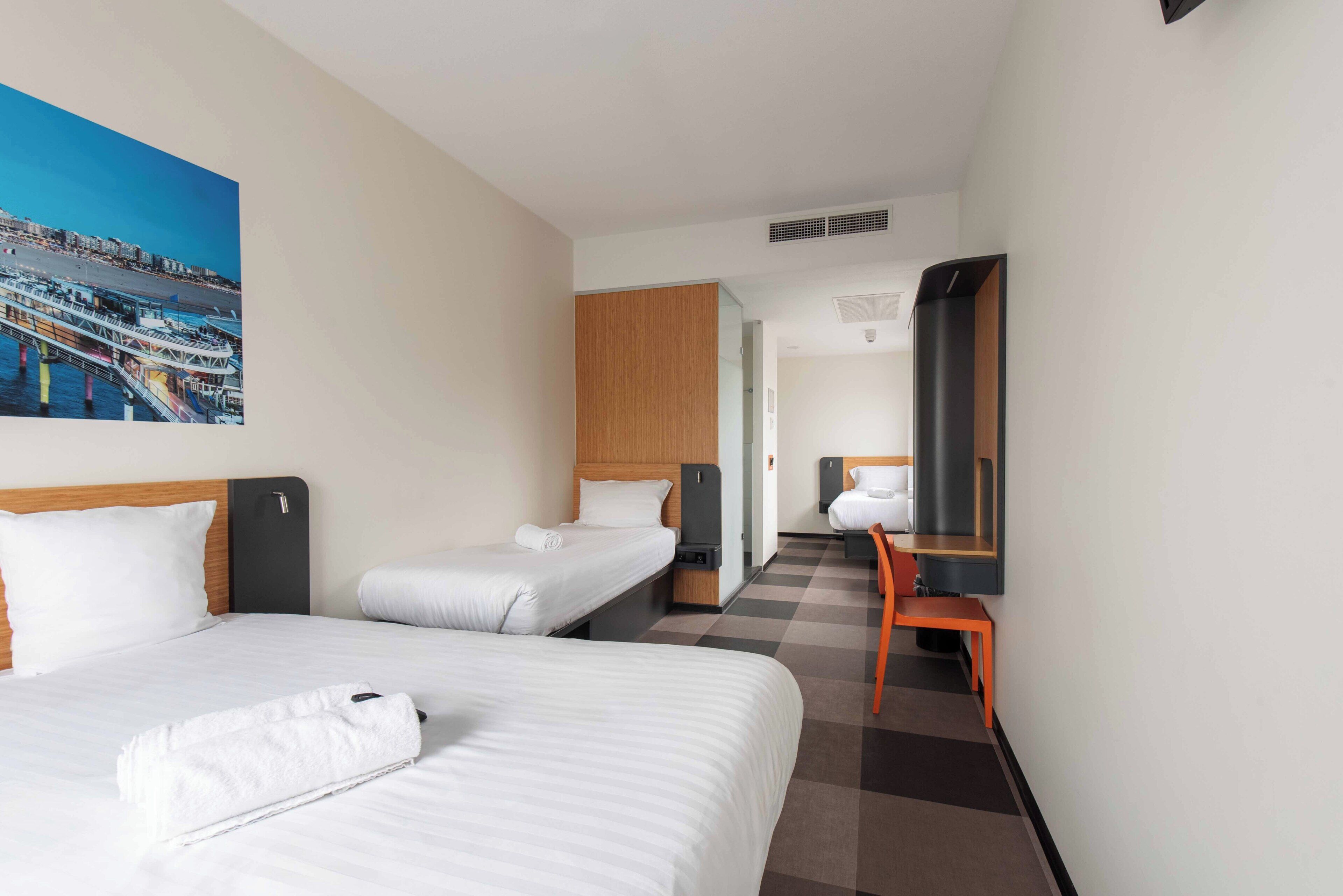 Habitación de hotel con dos camas, un escritorio, una silla, un ventanal grande y un cuadro en la pared.