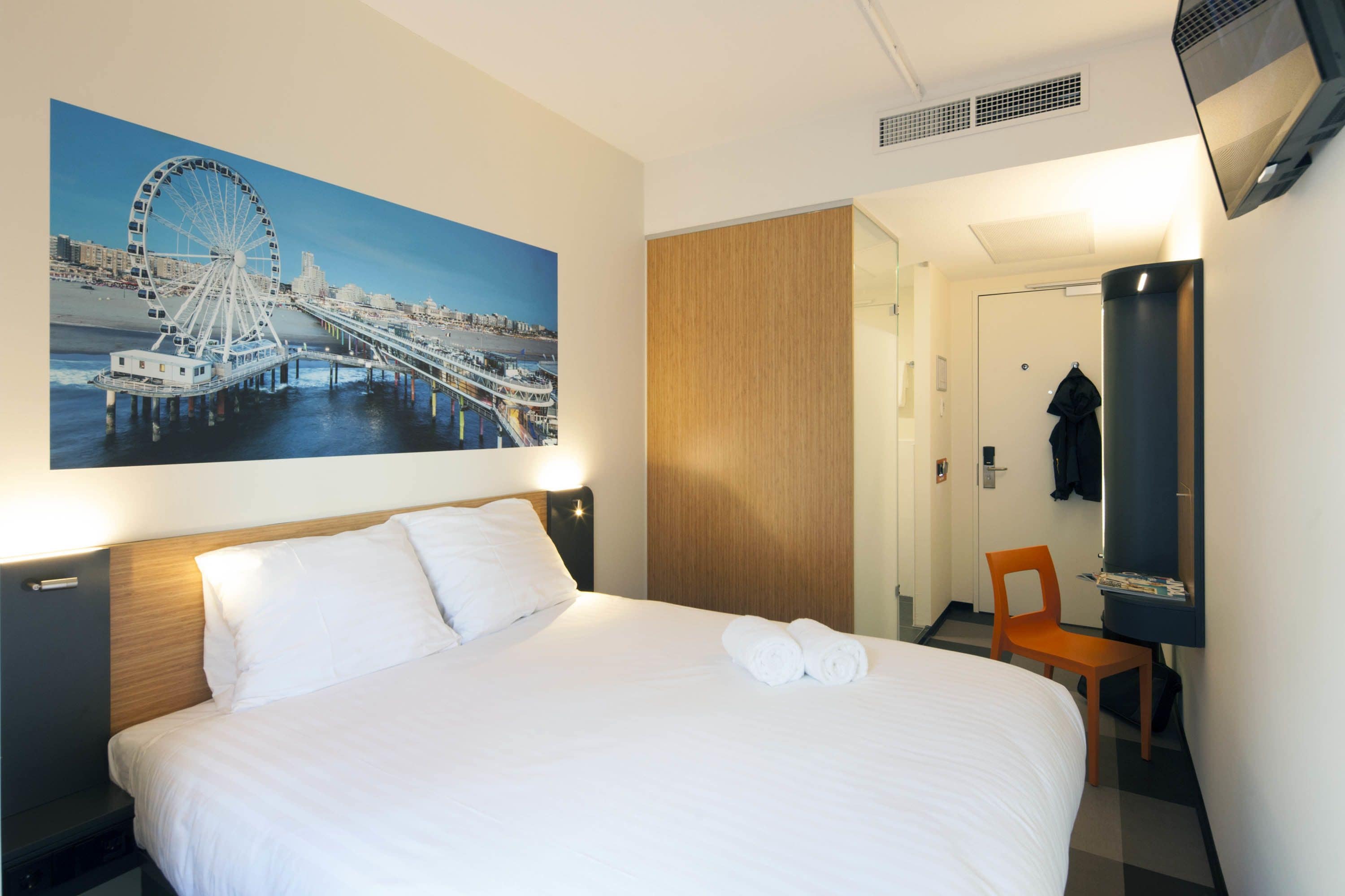 A hotel room with a large bed, a wooden headboard, a wall-mounted TV, a wooden door, and a chair.