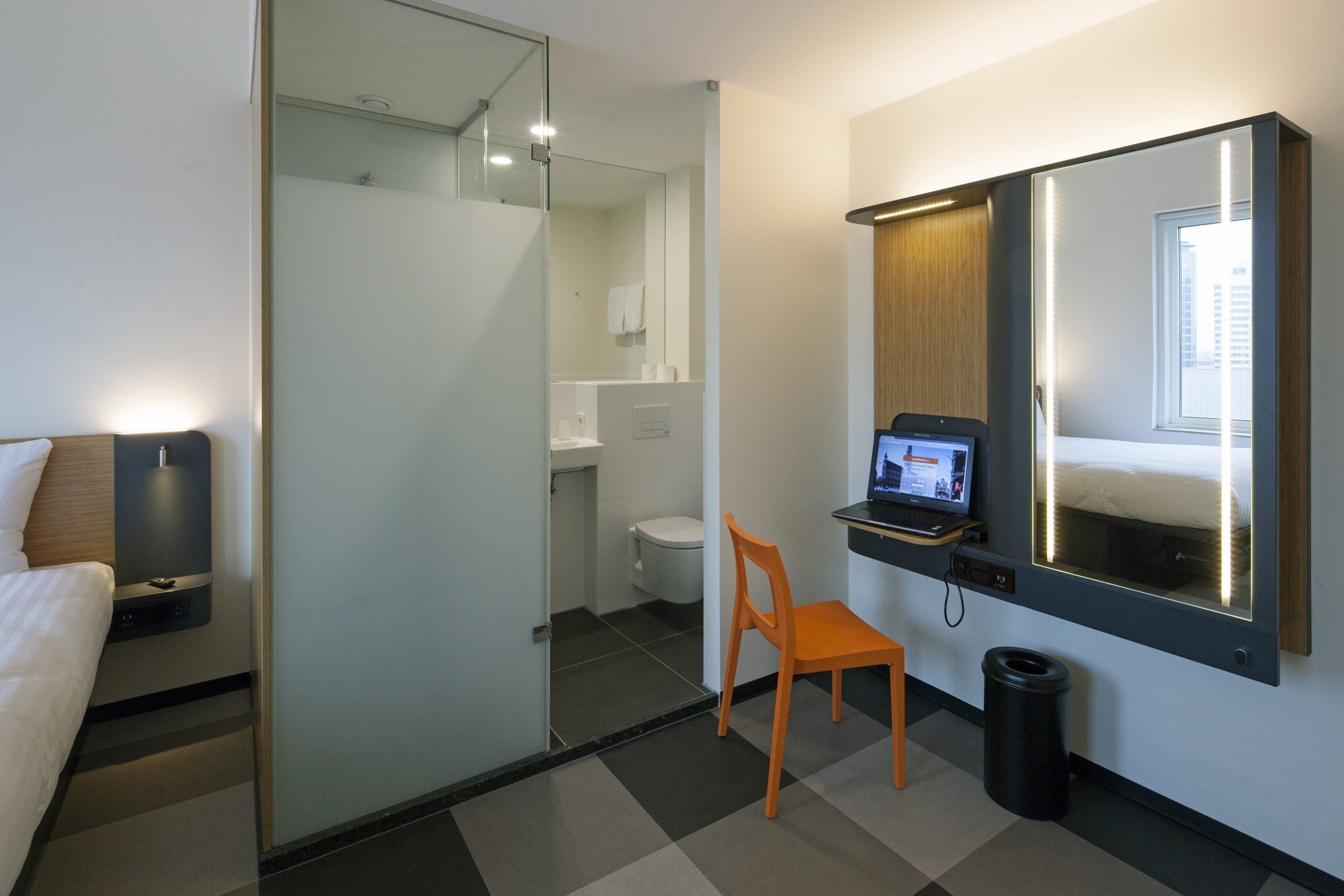 A hotel room with a bed, a desk with a laptop, a chair, a trash bin, and a glass partition separating the room from the bathroom.