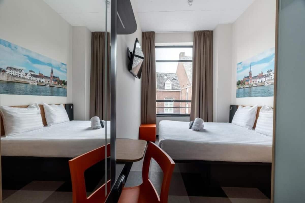 Een hotelkamer met twee bedden, een bureau, stoelen en uitzicht op een stadsbeeld door het raam.