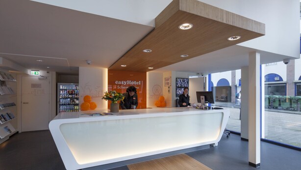 Een moderne receptiehal van een hotel met een wit receptiebalie, een potplant en een bordje met 'easyHotel Scheveningen'.
