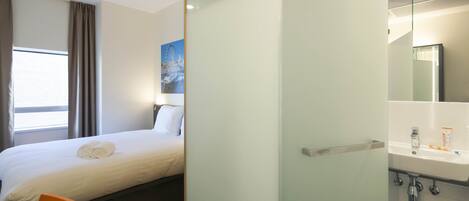 A hotel room with a bed, a chair, a bathroom with a sink, and a large window with curtains.