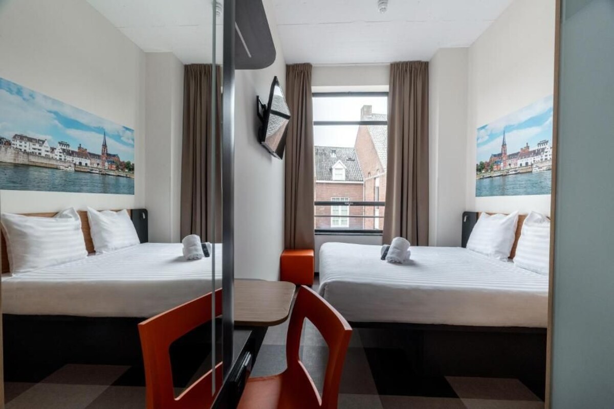 Quarto de hotel com duas camas, uma escrivaninha, cadeiras e vista para a cidade pela janela.