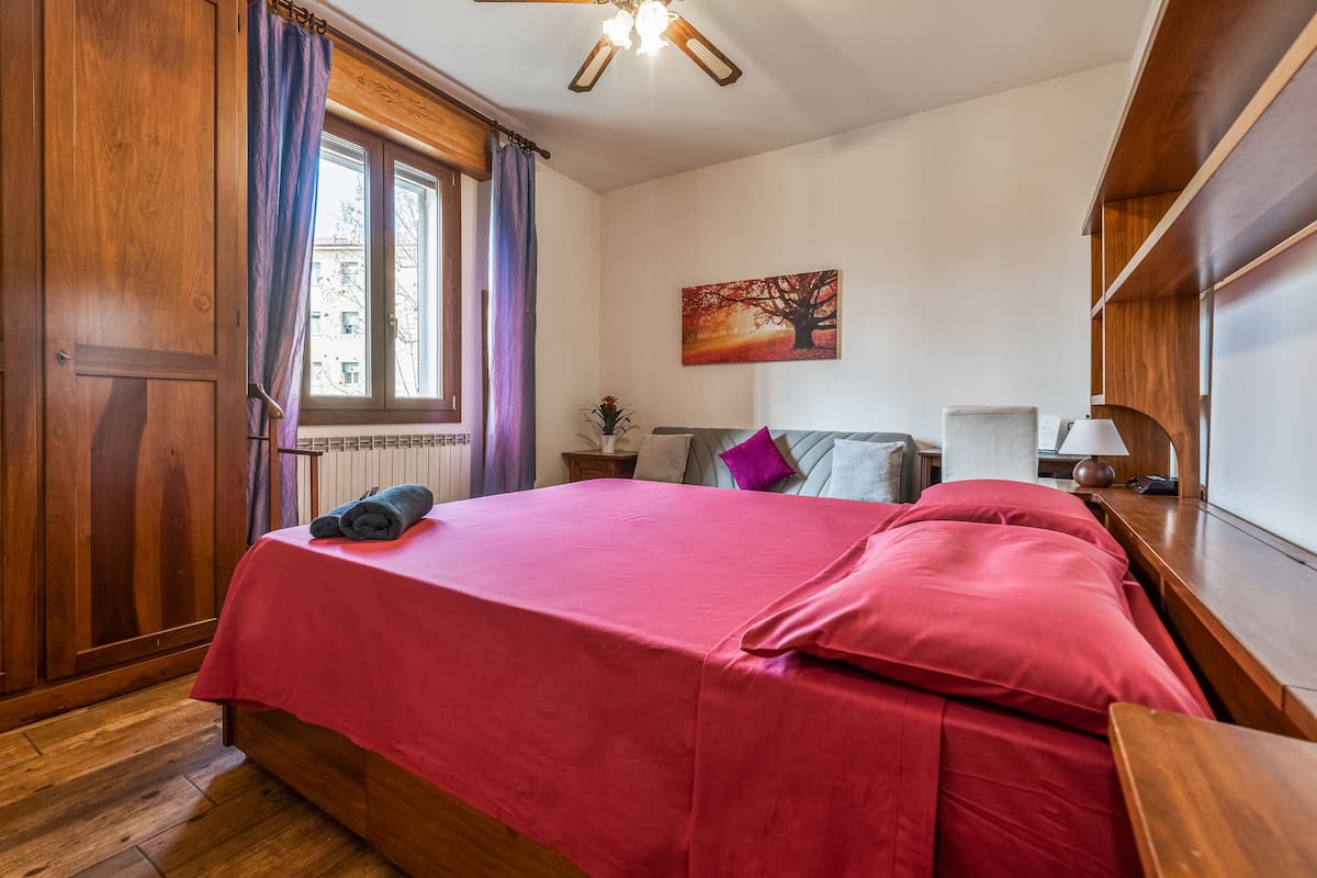 A bedroom with a wooden bed, a window with curtains, a nightstand with a lamp, and a painting on the wall.