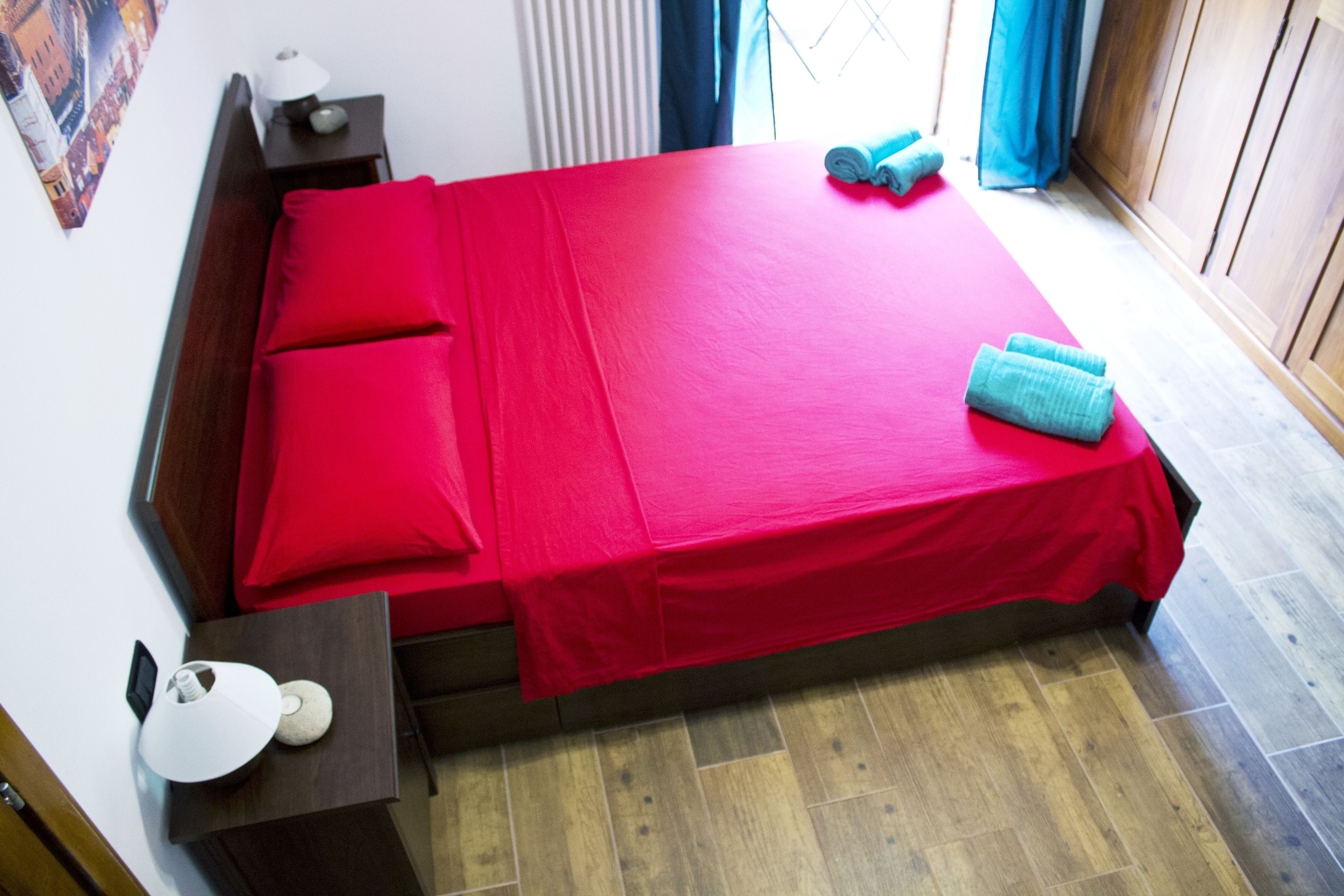 Een net opgemaakt bed met rode beddengoed, een houten hoofdbord, een nachtkastje met een lamp en een raam met gordijnen.