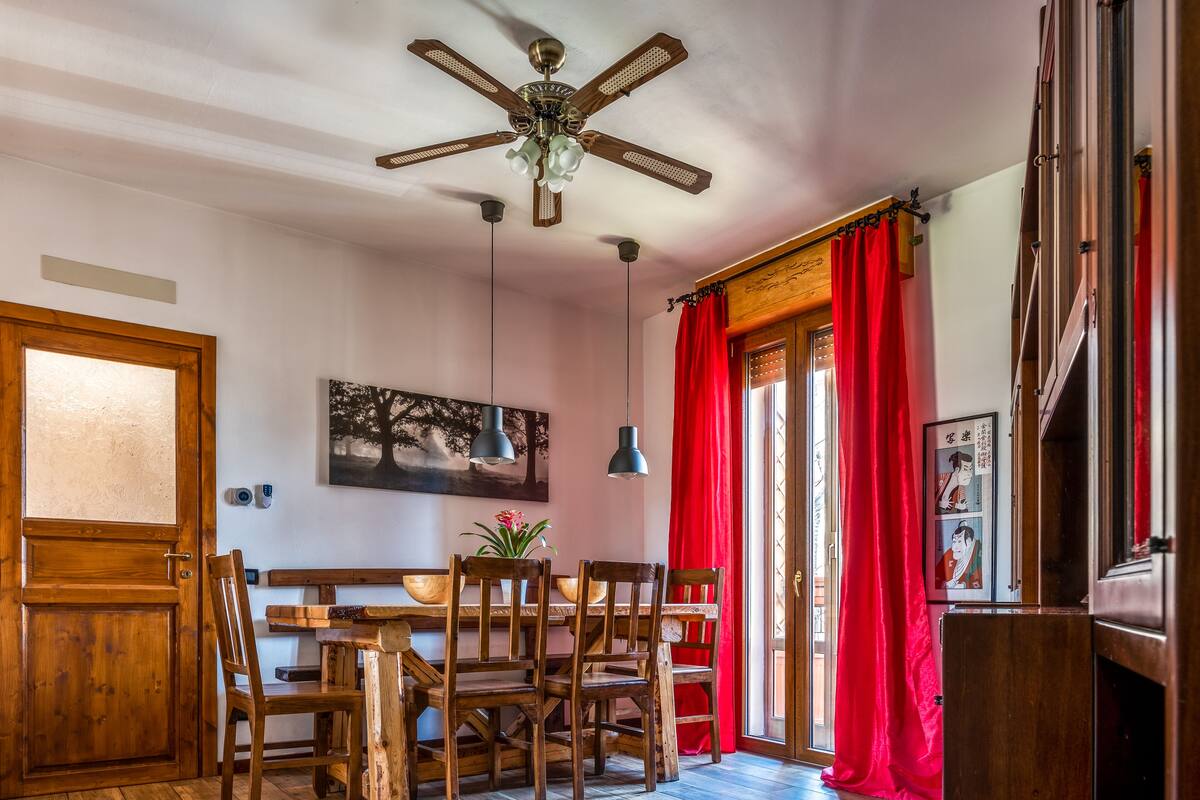 A dining area with a wooden table and chairs, a ceiling fan, pendant lights, and red curtains.