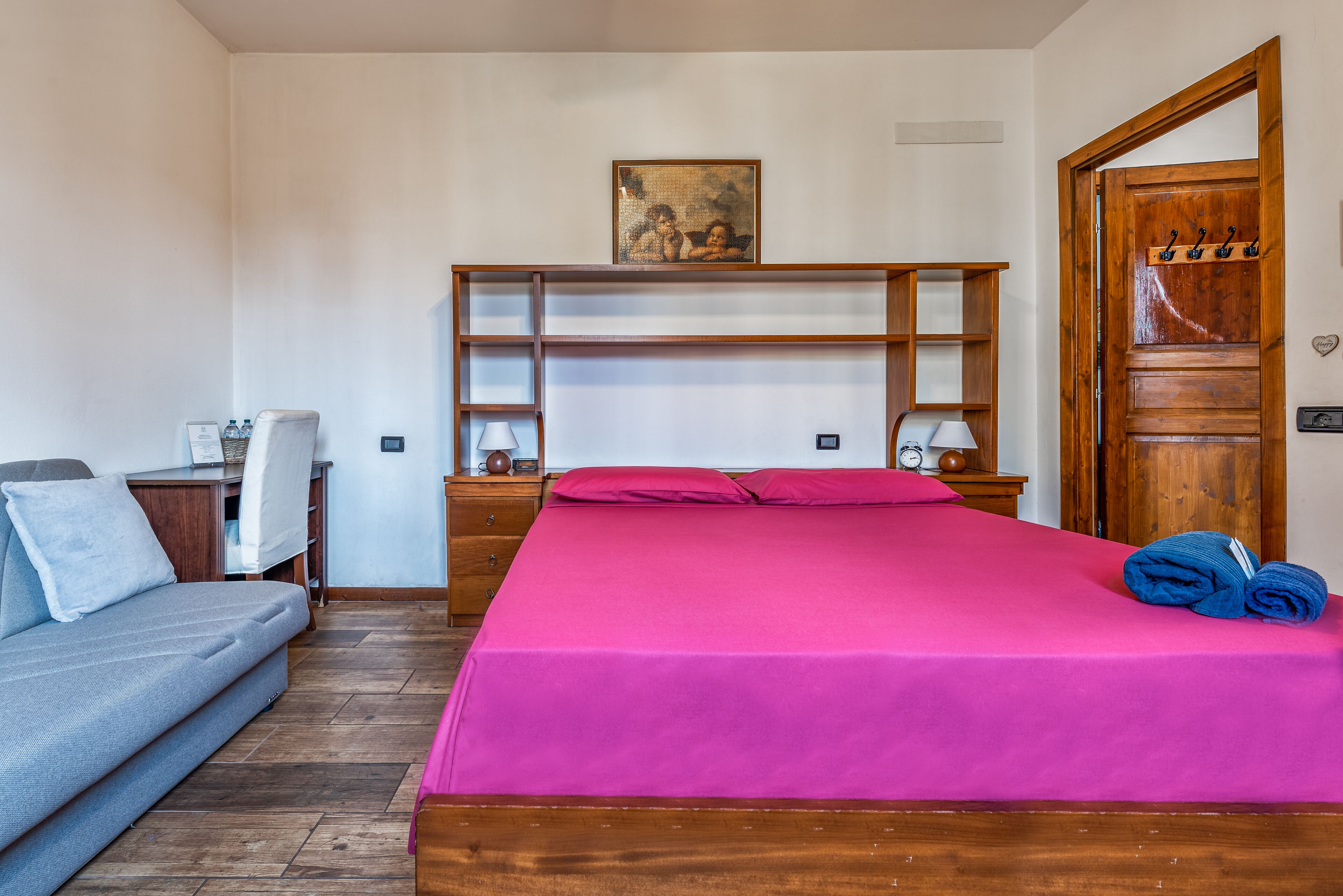 A bedroom with a bed, a sofa, a wooden wardrobe, and a door with hooks.
