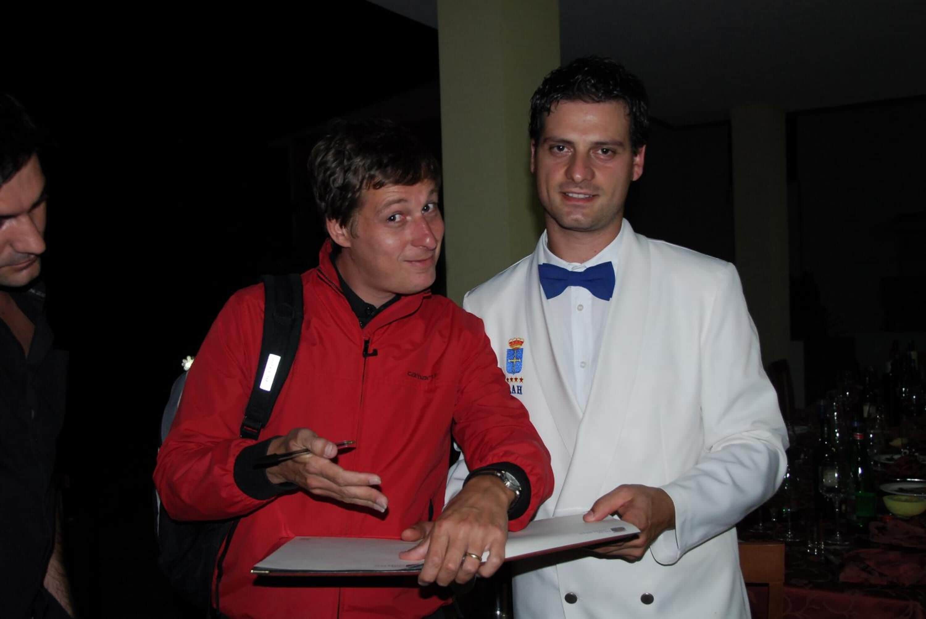 Un hombre con una chaqueta roja sosteniendo una tableta, de pie junto a un hombre con traje blanco y una corbata azul.