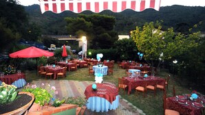 An outdoor dining area with round tables covered in red tablecloths, surrounded by chairs, and red umbrellas providing shade.
