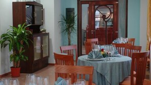 A dining area with a round table set for four, surrounded by wooden chairs. A potted plant is on the left, and a cabinet with decorative items is against the wall.
