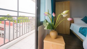 A hotel room with a balcony, a wooden nightstand with a vase of flowers, a bed with a blue blanket, and a wooden wardrobe. - Hanoi Prime Center Hotel (Hanoi)