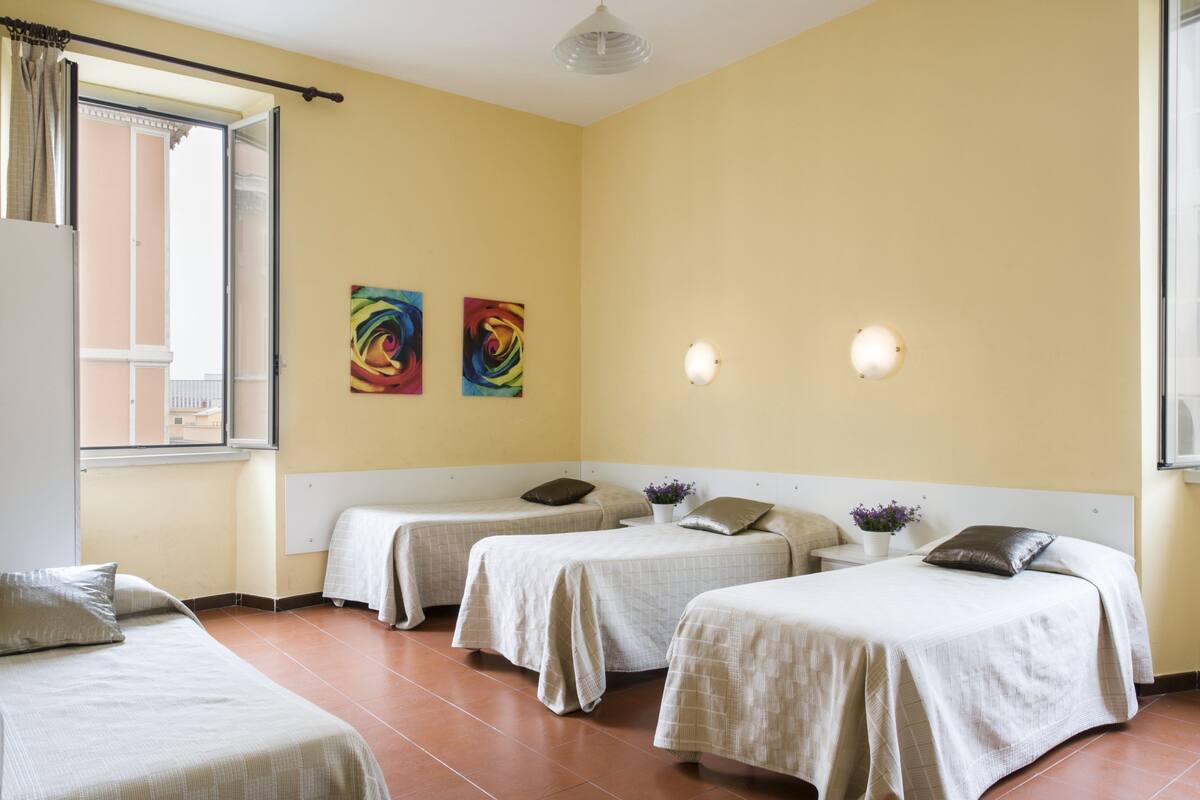A hotel room with two single beds, a small table, two wall-mounted lights, and two colorful paintings on the wall.