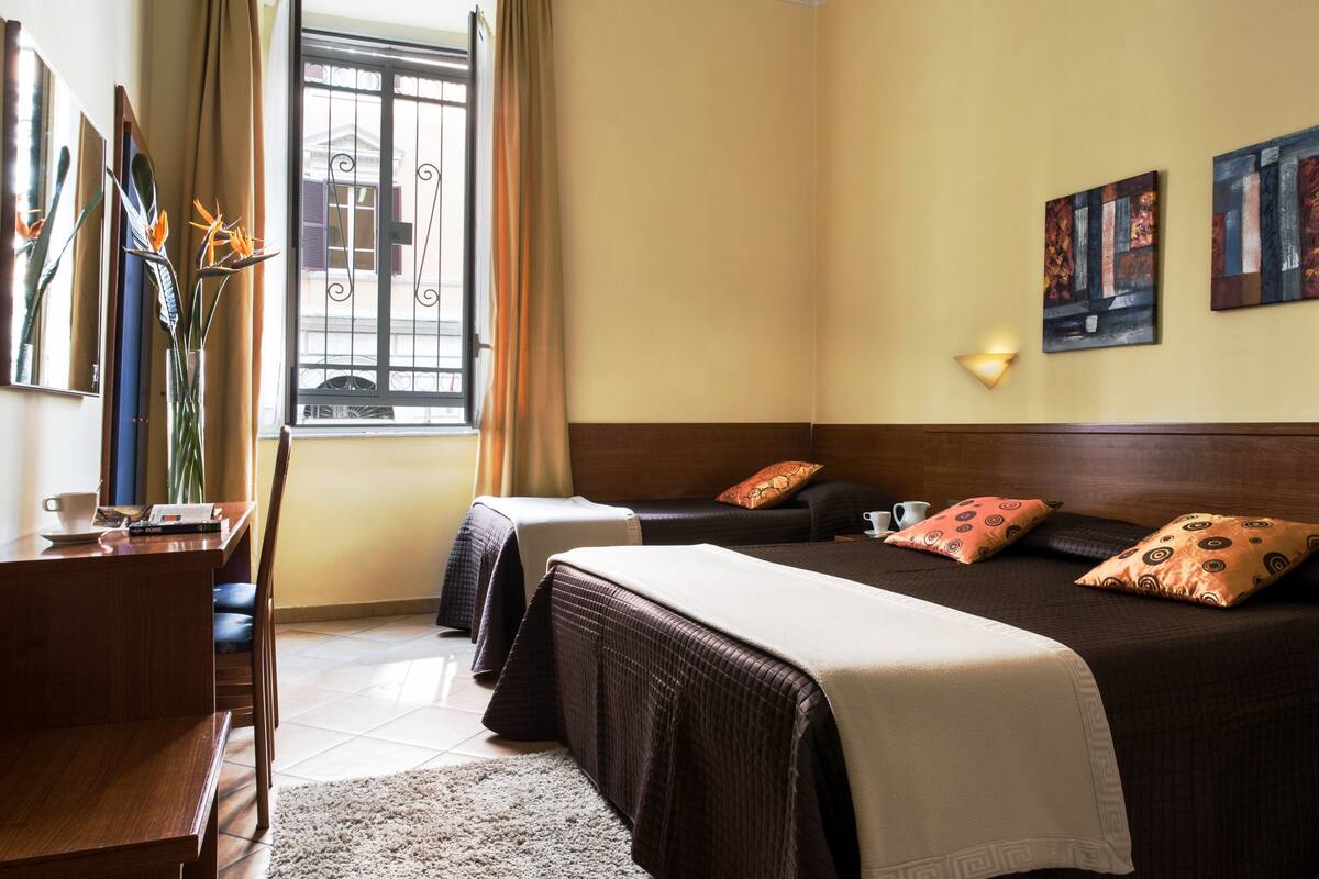 A hotel room with two beds, a desk, a chair, a window with curtains, and artwork on the wall.