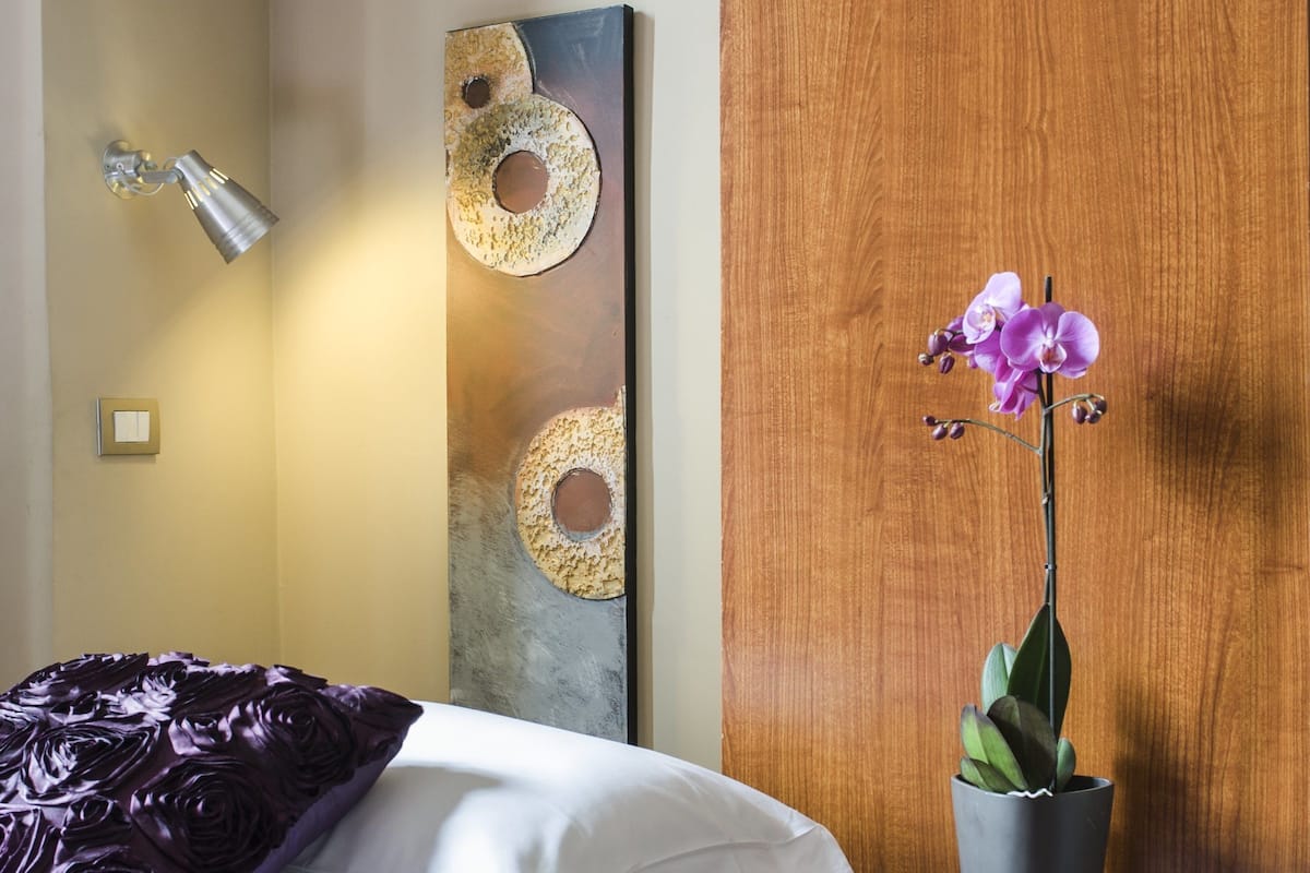 A neatly made bed with a purple blanket, a bedside table with a magazine, a potted orchid, and a wall with circular artwork and a light fixture.