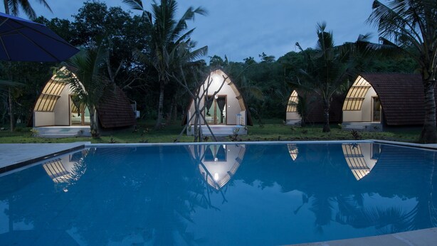 A pool area with a clear blue water surface, surrounded by palm trees and a few small, dome-shaped structures with open doors. - Apit Lawang Villas (Penida Island)