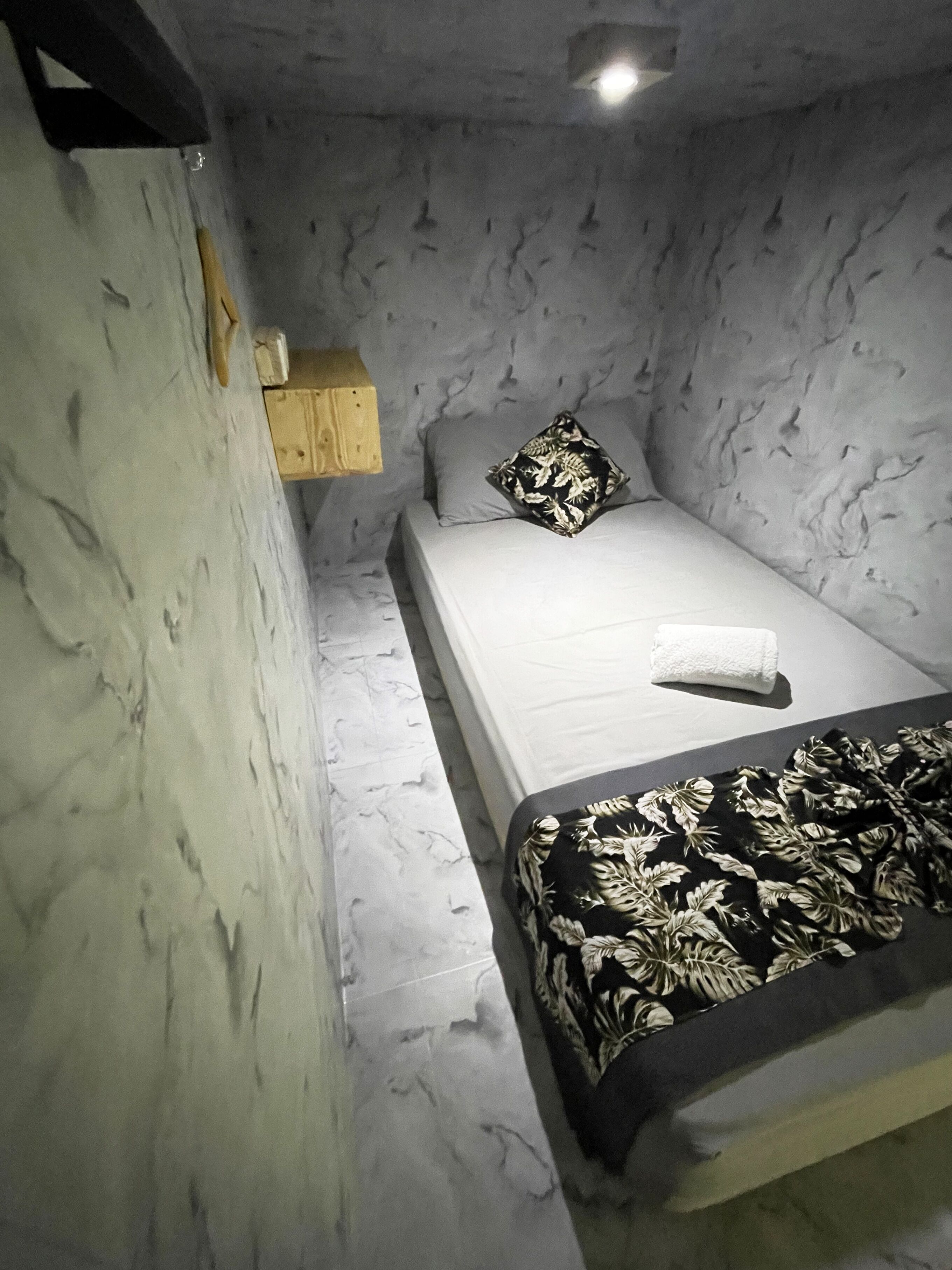Una habitación pequeña y tenuemente iluminada, con una cama individual, una mesita de noche de madera y un estante en la pared.