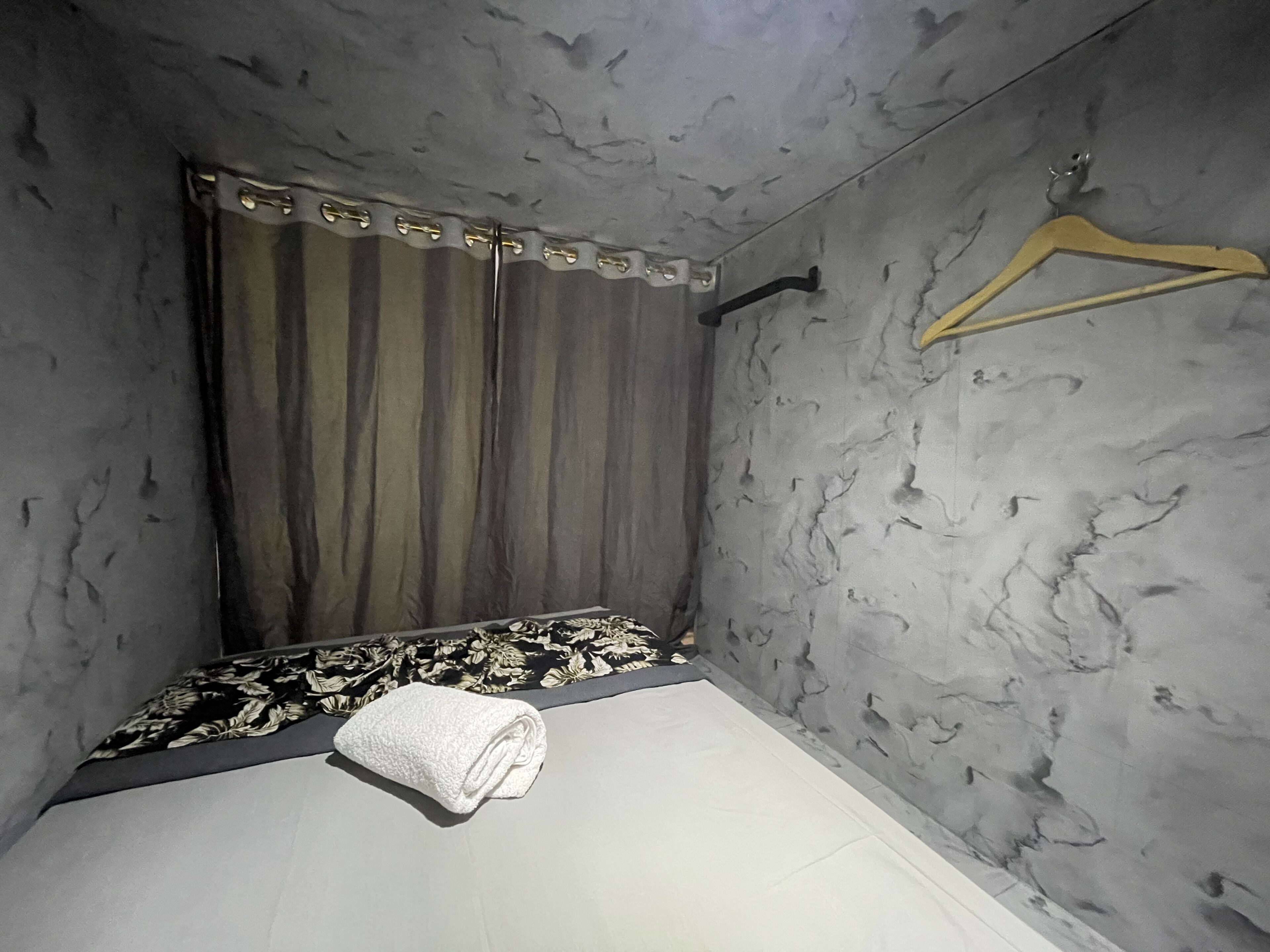 Une petite pièce faiblement éclairée, avec un lit, un rideau à rayures et un cintre en bois accroché au mur.