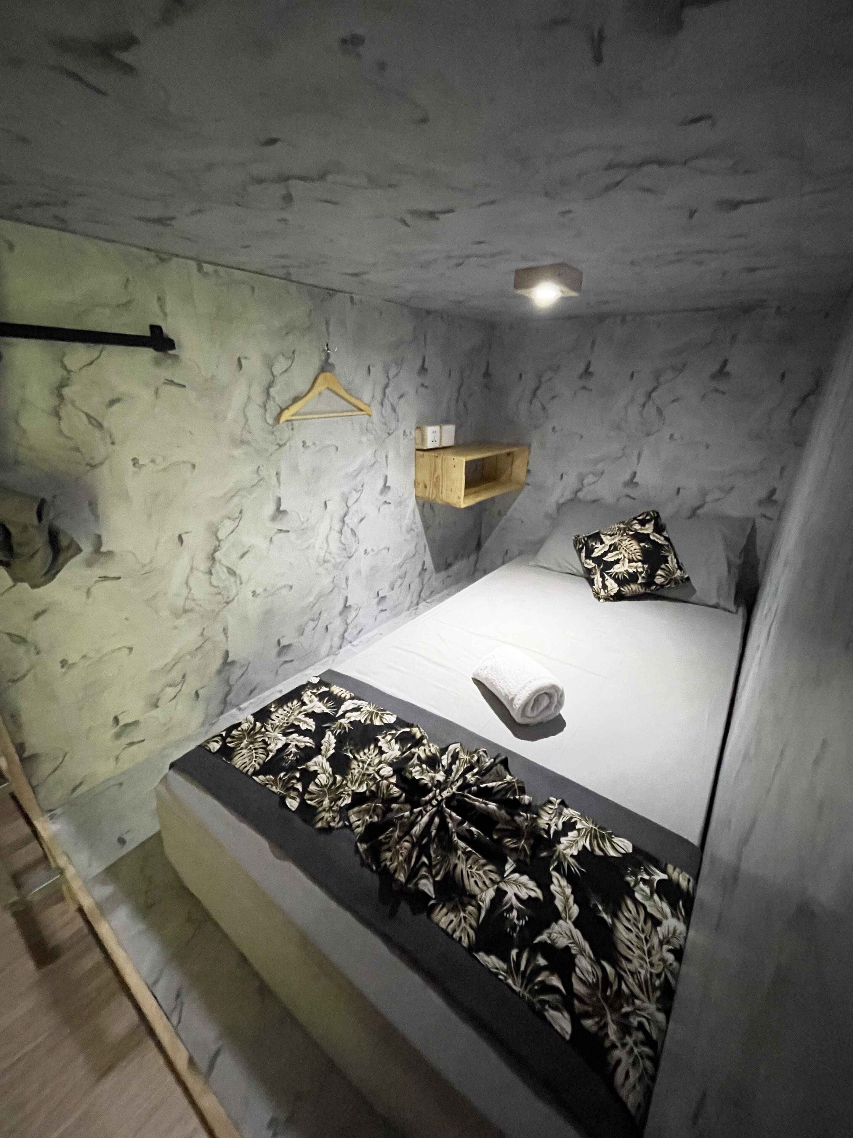 A bedroom with a bed, a hanging light, a wooden shelf, and a patterned bedspread.