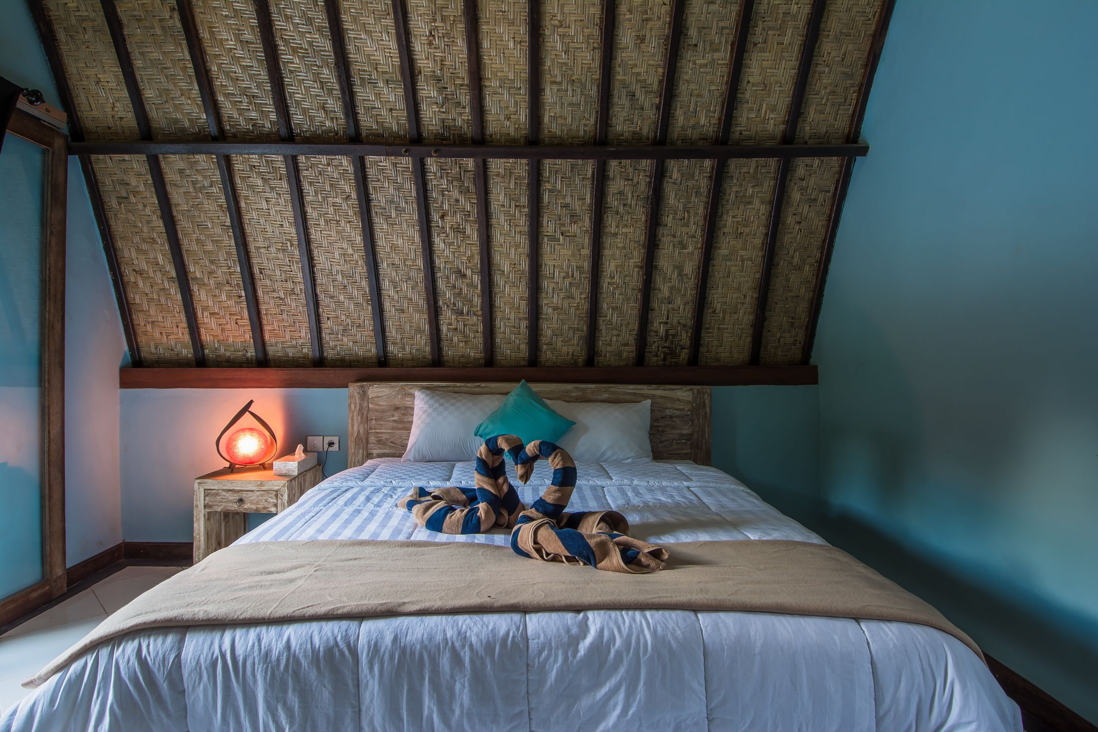 A bed with a woven ceiling, a bedside table with a lamp, and a blue pillow.