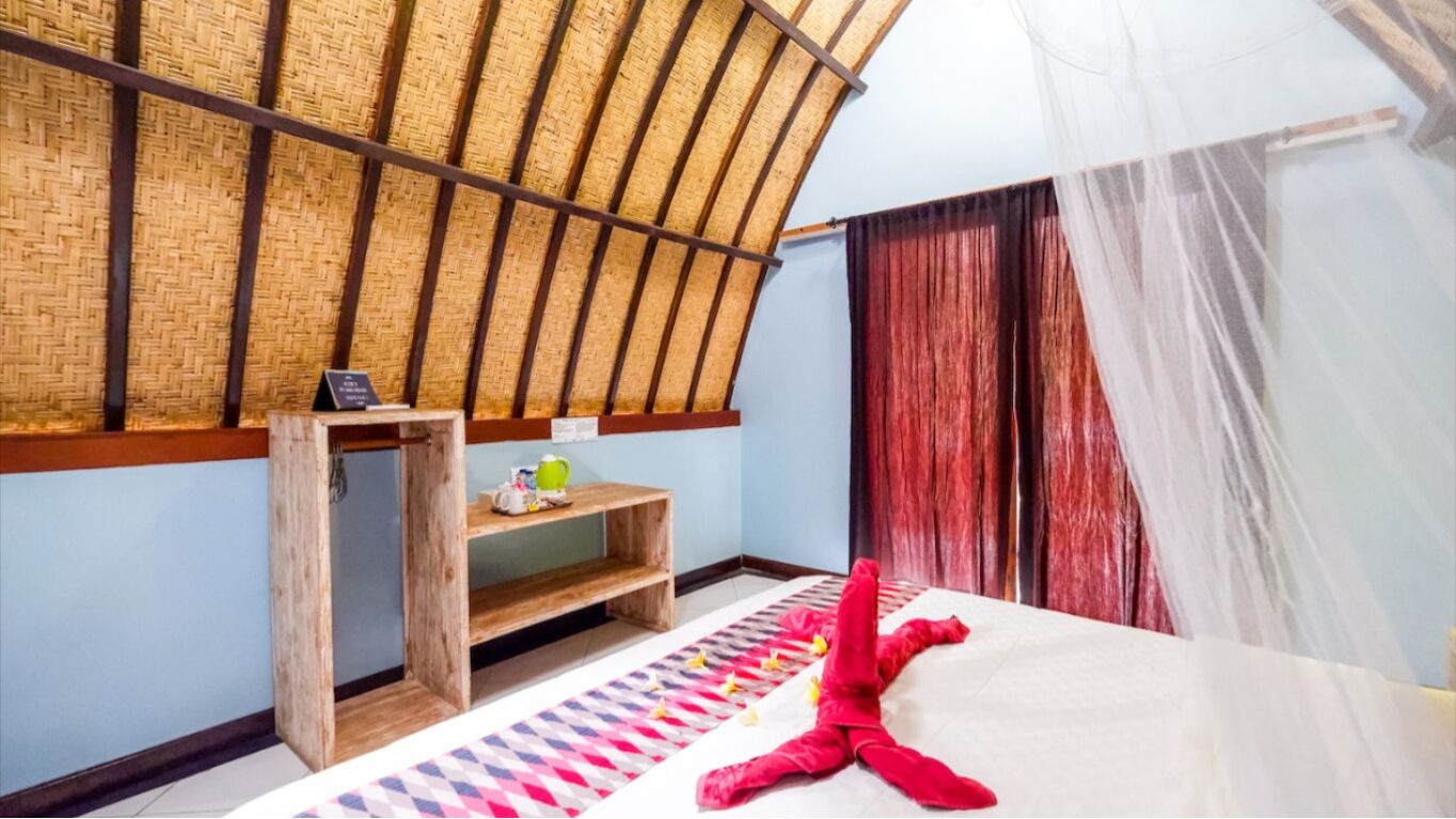 Una habitación con techo de paja, una cama con un peluche rojo, una estantería con una tetera y una cortina con mosquitero.