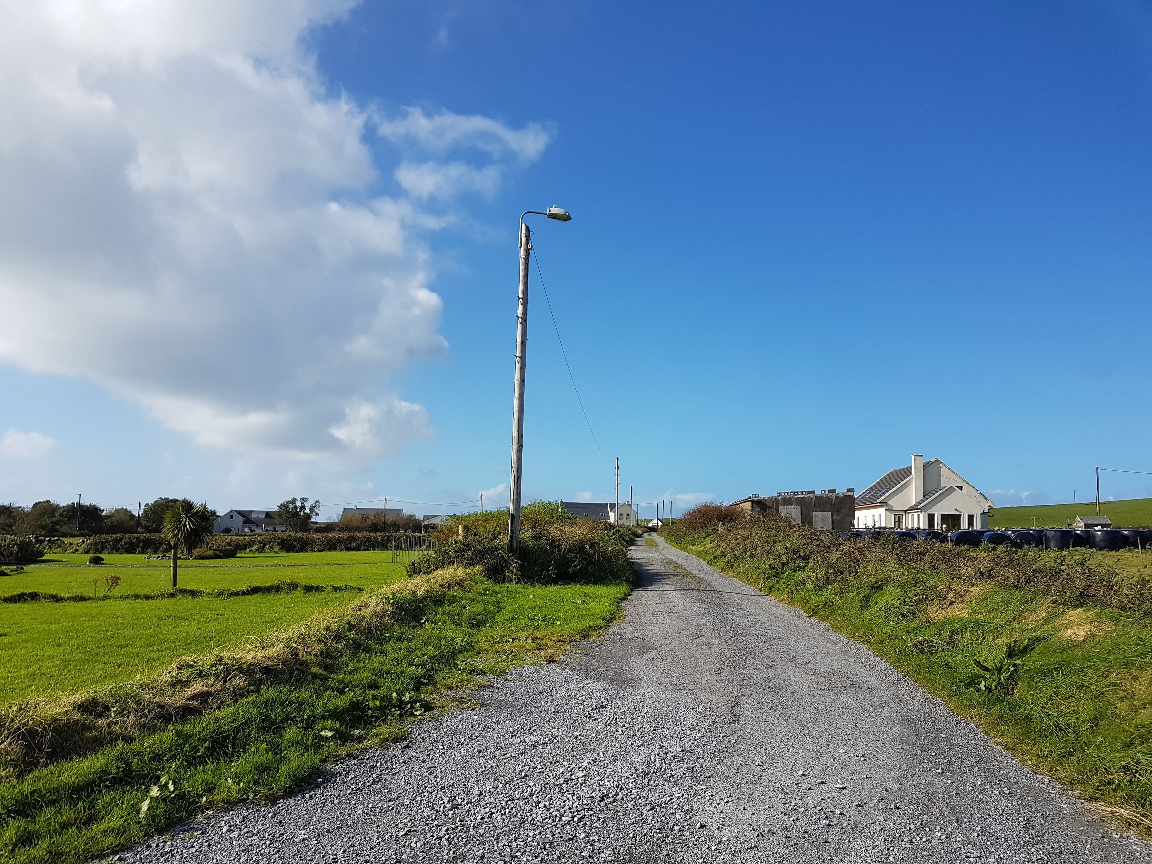 A rural road with a gravel surface, a street lamp, and a white building with a sloped roof in the distance.